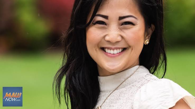 A smiling woman with long dark hair wearing a white textured top, photographed outdoors against a blurred green background. A small 'MAUI NOW.